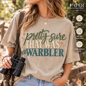 May include: A beige short-sleeve t-shirt featuring the green and blue text "pretty sure THAT WAS A WARBLER" with small star and checkered accents. The shirt is worn with blue denim jeans and a pair of black binoculars held in the hand.