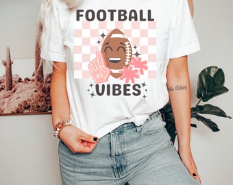 May include: White t-shirt with the text "FOOTBALL VIBES" in black. A pink and white checkered background with a cartoon football with a smiling face and pink flowers.