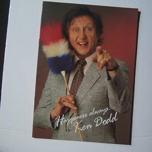 May include: A promotional image of Ken Dodd, wearing a grey suit and pointing. He holds a feather duster with red, white, and blue feathers. The image includes the text "Happiness always... Ken Dodd".