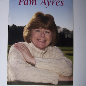 May include: A book cover with the title "Pam Ayres" in red lettering. The image shows a woman with brown hair wearing a cream-colored knitted sweater. She is smiling and has her arms crossed. The background is a blue sky and a landscape.