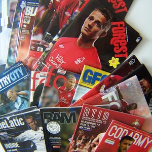 May include: A pile of football matchday programmes, predominantly featuring players in red and white kits. The covers display team names such as "Forest" and "Derby County", along with player images and match information.