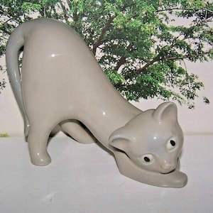 May include: A ceramic figurine of a grey cat in a crouching position. The cat has large, black eyes and a long, curled tail.