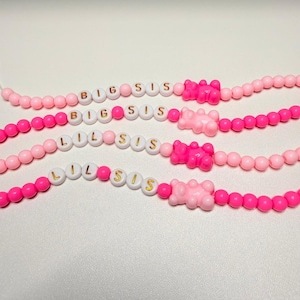 May include: Four beaded bracelets in shades of pink. The bracelets have white letter beads spelling "BIG SIS" and "LIL SIS" with a pink gummy bear charm. The beads are strung on a white cord.