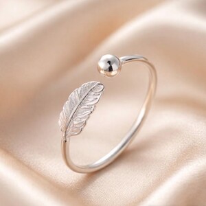 May include: A silver ring featuring a feather motif and a small ball. The open design allows for size adjustment. The feather is finely detailed. The ring is displayed against a soft, neutral-toned background.