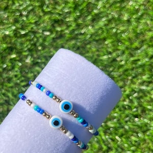 May include: Two beaded bracelets with gold accents and blue evil eye charms. The bracelets are on a white surface.