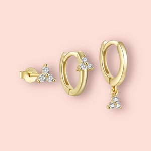 May include: A set of three gold earrings featuring small, clear cubic zirconia stones. The earrings are shaped like a triangle and are designed for earlobe wear.