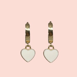 Puede incluir: Un par de pendientes de aro dorados con un dije de corazón blanco que cuelga de cada aro.