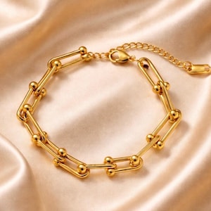 Puede incluir: Una pulsera dorada con un diseño de eslabones en forma de U y pequeños detalles de bolas. La pulsera es de metal brillante y tiene un cierre de cadena ajustable. La joya se muestra sobre una superficie satinada.