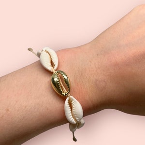 Puede incluir: Un brazalete de conchas blancas con un acento dorado. El brazalete está hecho de conchas naturales y una cuerda fina.