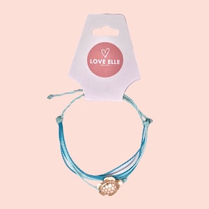 Op de afbeelding: Een lichtblauwe en witte vriendschapsband met een gouden schildpadbedel. De armband is met een knoop vastgemaakt en heeft een label met de tekst "LOVE ELLE JEWELLERY".