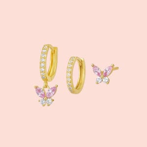 Puede incluir: Un conjunto de tres pendientes dorados. Un par son pendientes de aro con piedras transparentes y un dije de mariposa con piedras rosas y blancas. El segundo es un solo pendiente de aro con piedras transparentes. El tercero es un pendiente de botón de mariposa con piedras rosas y blancas.