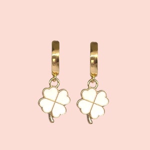 Peut inclure: Une paire de boucles d'oreilles créoles dorées avec un charme trèfle à quatre feuilles blanc suspendu à chaque créole.