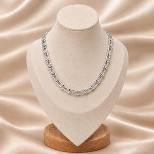 May include: A silver-toned necklace featuring a series of interconnected U-shaped links, each accented with small spherical beads at the joints. The chain is displayed on a neutral-coloured bust, showcasing a uniform metallic finish and a structured design.
