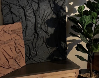 May include: A black and brown abstract art piece hangs on a wall above a wooden bench with a brown top. A potted plant with large green leaves sits on the floor next to the bench.