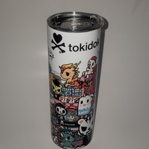 Pode incluir: Um copo de aço inoxidável branco com um logotipo preto e branco de caveira e ossos cruzados que diz "tokidoki". O copo é coberto com um padrão colorido de personagens de desenhos animados e designs.