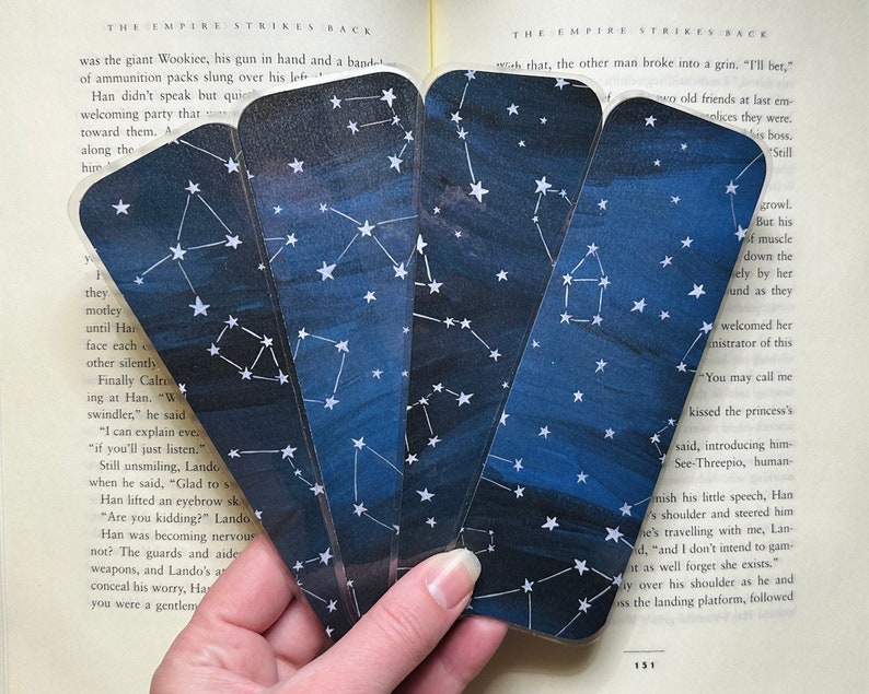 Four bookmarks are held up in front of the pages of an open book. All of the bookmarks have a dark blue varied background with white stars and constellations.