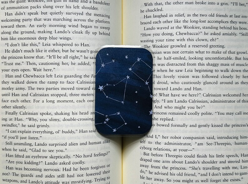A mini bookmark is laid out on the pages of an open book. It has a dark blue varied background with white stars and constellations.