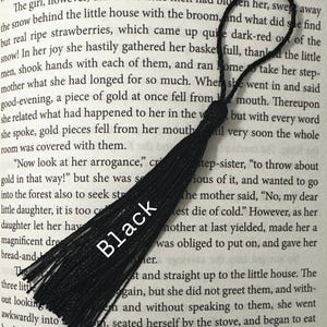 May include: A black tassel, likely for a bookmark or decoration, lies diagonally across a book page. The tassel is made of multiple strands and has a loop at the top. The word "Black" is printed on the tassel.