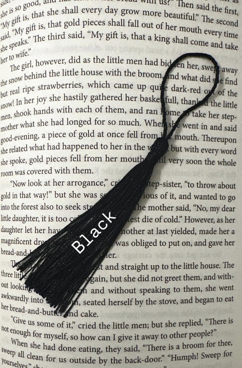May include: A black tassel, likely for a bookmark or decoration, lies diagonally across a book page. The tassel is made of multiple strands and has a loop at the top. The word "Black" is printed on the tassel.