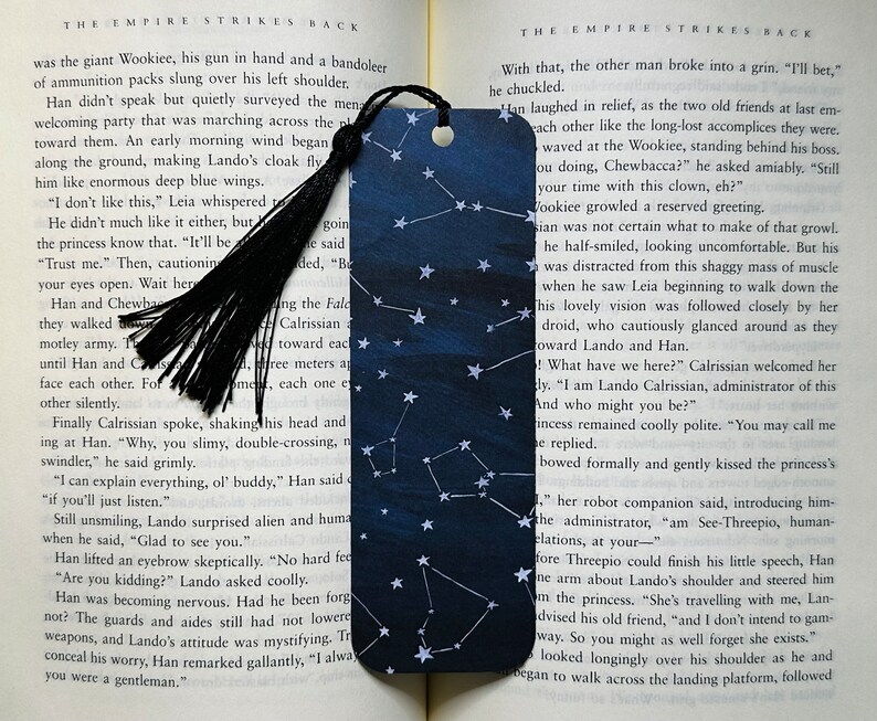 A bookmark is laid out on the pages of an open book. It has a dark blue varied background with white stars and constellations. The bookmark has a black tassel.