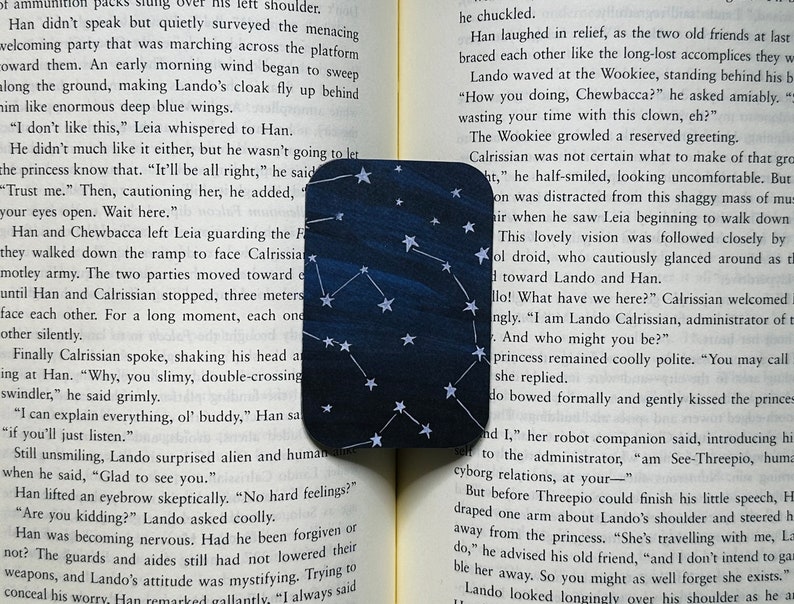 A mini bookmark is laid out on the pages of an open book. It has a dark blue varied background with white stars and constellations.