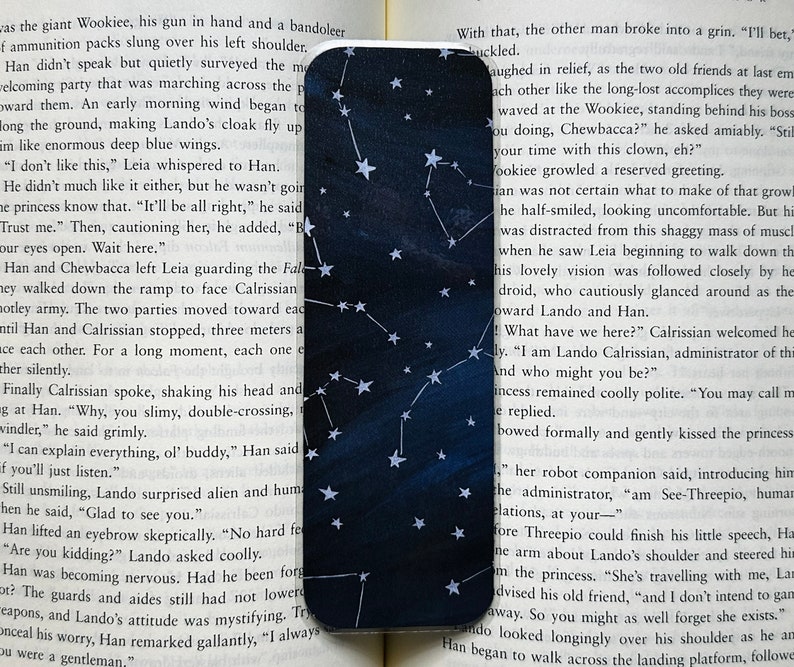 A bookmark is laid out on the pages of an open book. It has a dark blue varied background with white stars and constellations.