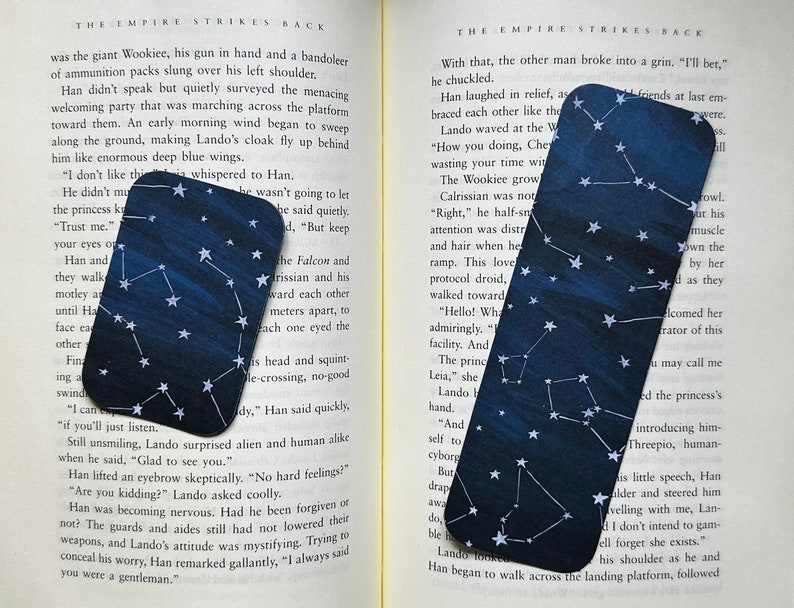 Two bookmarks are laid out on the pages of an open book. They both have a dark blue varied background with white stars and constellations.