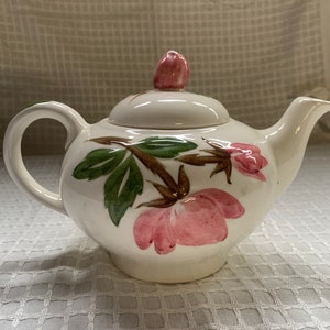 Könnte beinhalten: Eine weiße Teekanne aus Keramik mit einem rosafarbenen Rosenmuster. Die Teekanne hat einen Deckel mit einem rosafarbenen Rosenknopf. Die Teekanne steht auf einer weißen Tischdecke.