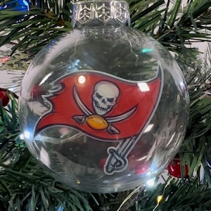 May include: Clear glass Christmas ornament featuring the Tampa Bay Buccaneers logo. The logo is a red flag with a skull and crossed swords. The ornament hangs on a green Christmas tree with white lights.