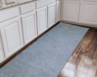 May include: A light blue patterned kitchen runner rug with a geometric design. The rug is placed in a kitchen with white cabinets and hardwood floors.