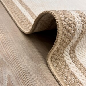 May include: Close-up of a beige and cream-coloured woven rug with a striped design. The rug has a rolled edge, revealing the texture and construction of the material. The rug is placed on a light-coloured wooden floor.