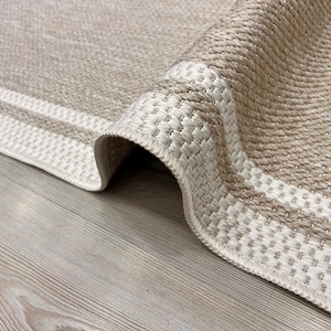 Puede incluir: Primer plano de una alfombra beige tejida con un borde enrollado. La alfombra presenta un patrón texturizado con un borde más claro. La paleta de colores neutros y la textura tejida sugieren un artículo de decoración del hogar.