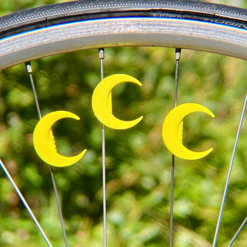 May include: Close-up of a bicycle wheel with three bright yellow crescent-shaped objects attached to the spokes. The objects are evenly spaced and stand out against the silver wheel and green background.