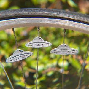May include: Close-up of a bicycle wheel with three small, gray, UFO-shaped ornaments hanging from the spokes. The ornaments have a classic flying saucer design. The background is blurred green foliage.