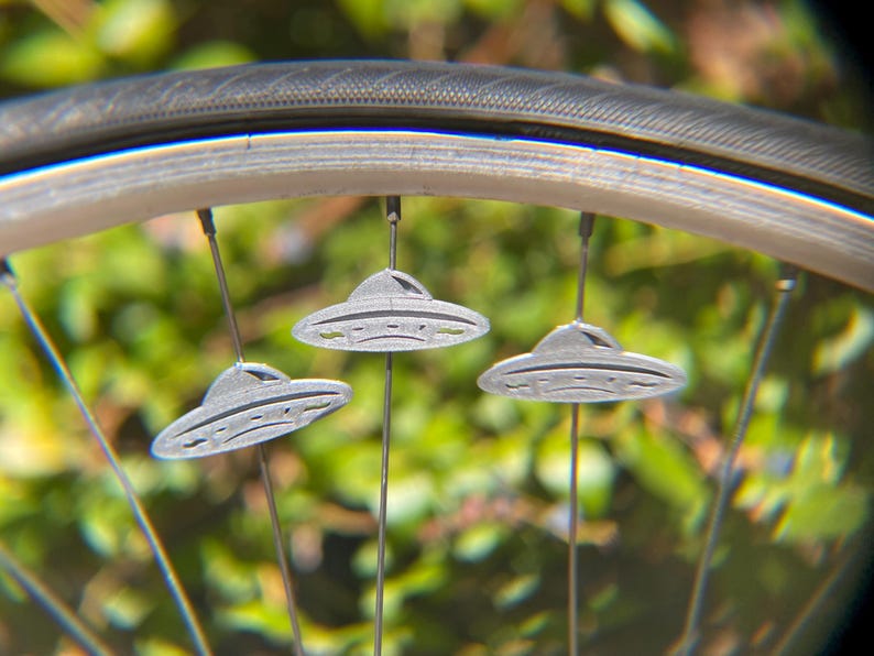 May include: Close-up of a bicycle wheel with three small, gray, UFO-shaped ornaments hanging from the spokes. The ornaments have a classic flying saucer design. The background is blurred green foliage.