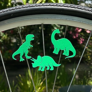 May include: Green dinosaur-shaped bicycle wheel decorations. The image shows three different dinosaur silhouettes attached to the spokes of a bicycle wheel. The dinosaurs are bright green and appear to be made of a flat material.