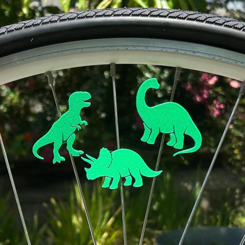 May include: Green dinosaur-shaped bicycle wheel decorations. The image shows three different dinosaur silhouettes attached to the spokes of a bicycle wheel. The dinosaurs are bright green and appear to be made of a flat material.