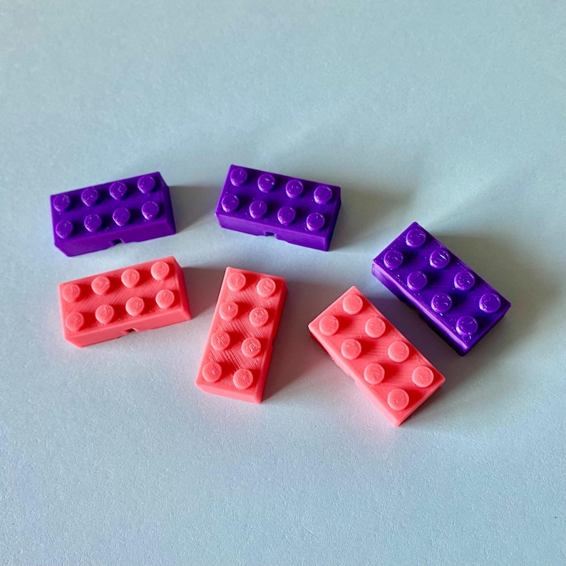 May include: Six colorful toy building blocks in shades of purple and coral pink are scattered on a light blue surface. Each block has a rectangular shape with circular studs on top, resembling classic construction toys.