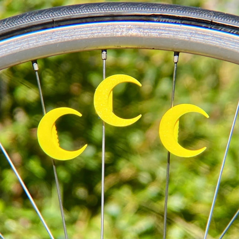 May include: Close-up of a bicycle wheel with three bright yellow crescent moon-shaped decorations attached to the spokes. The tire is black, and the wheel rim is silver. The background is a blurred green.