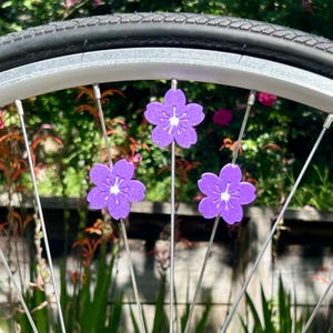 May include: Close-up of a bicycle wheel with three purple flower decorations attached to the spokes. The flowers have white centers and are set against a blurred green and pink background.