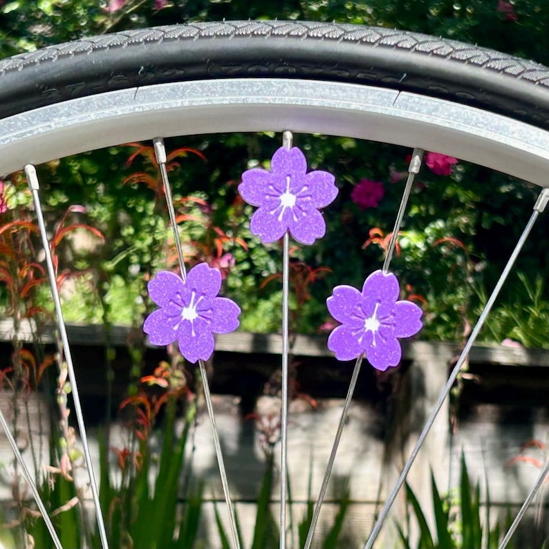 May include: Close-up of a bicycle wheel with three purple flower decorations attached to the spokes. The flowers have white centers and are set against a blurred green and pink background.