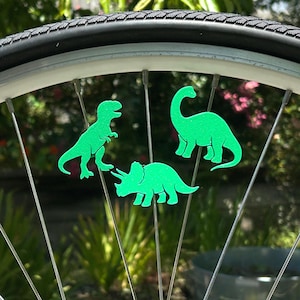 May include: A bicycle wheel with three bright green dinosaur-shaped reflectors attached to the spokes. The dinosaurs include a T-Rex, a Brontosaurus, and a Triceratops. The tire is black, and the background is blurred green foliage.