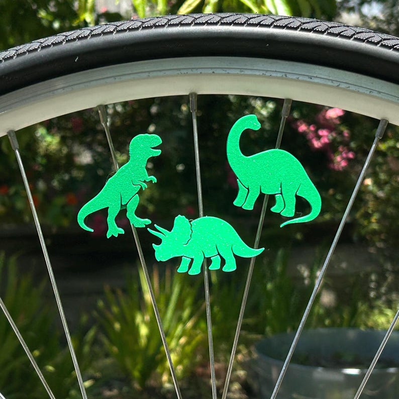May include: A bicycle wheel with three bright green dinosaur-shaped reflectors attached to the spokes. The dinosaurs include a T-Rex, a Brontosaurus, and a Triceratops. The tire is black, and the background is blurred green foliage.