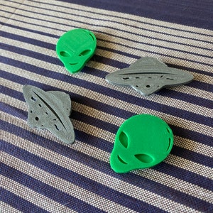 May include: Four 3D-printed alien and UFO-themed objects. Two green alien heads with large eyes and smiles, and two gray UFOs. The objects are on a blue and white striped surface.