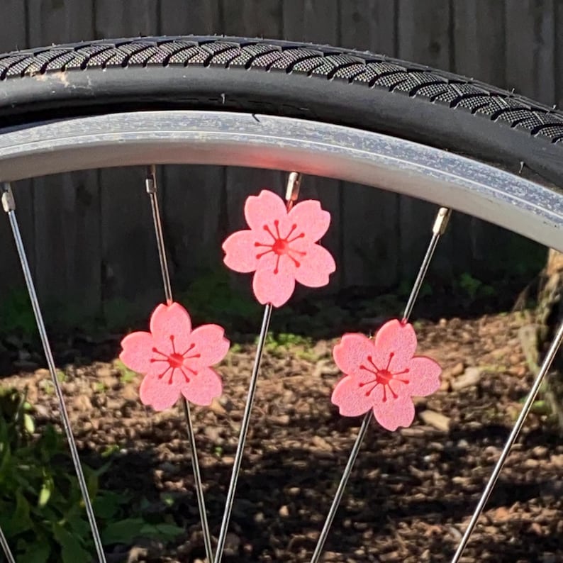 May include: Close-up of a bicycle wheel with three pink cherry blossom decorations attached to the spokes. The blossoms have a detailed center and are set against a blurred background of a tire and greenery.