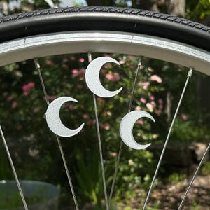 May include: Close-up of a bicycle wheel with three silver crescent moon charms attached to the spokes. The tire is black, and the wheel rim is silver. The background is blurred with green and pink foliage.