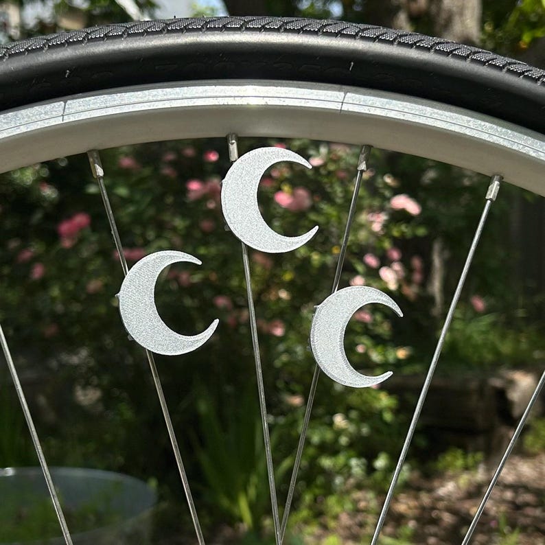 May include: Close-up of a bicycle wheel with three silver crescent moon charms attached to the spokes. The tire is black, and the wheel rim is silver. The background is blurred with green and pink foliage.