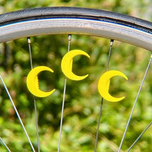 May include: Close-up of a bicycle wheel with three yellow crescent-shaped decorative accents attached to the spokes. The tire is black, and the wheel rim is silver. The background is a blurred green.