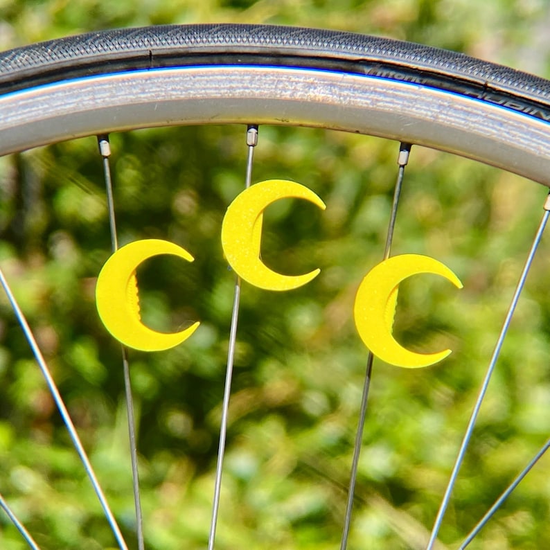 May include: Close-up of a bicycle wheel with three yellow crescent-shaped decorative accents attached to the spokes. The tire is black, and the wheel rim is silver. The background is a blurred green.