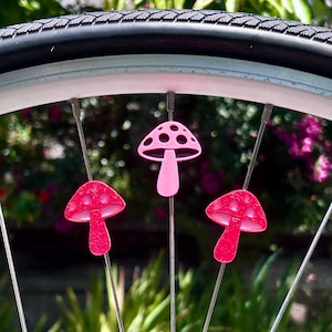 Puede incluir: Tres cuentas para radios en forma de seta de color rosa brillante fijadas a los radios met&aacute;licos de una rueda de bicicleta. Cada seta presenta una superficie texturizada y brillante con recortes circulares en el sombrero.
